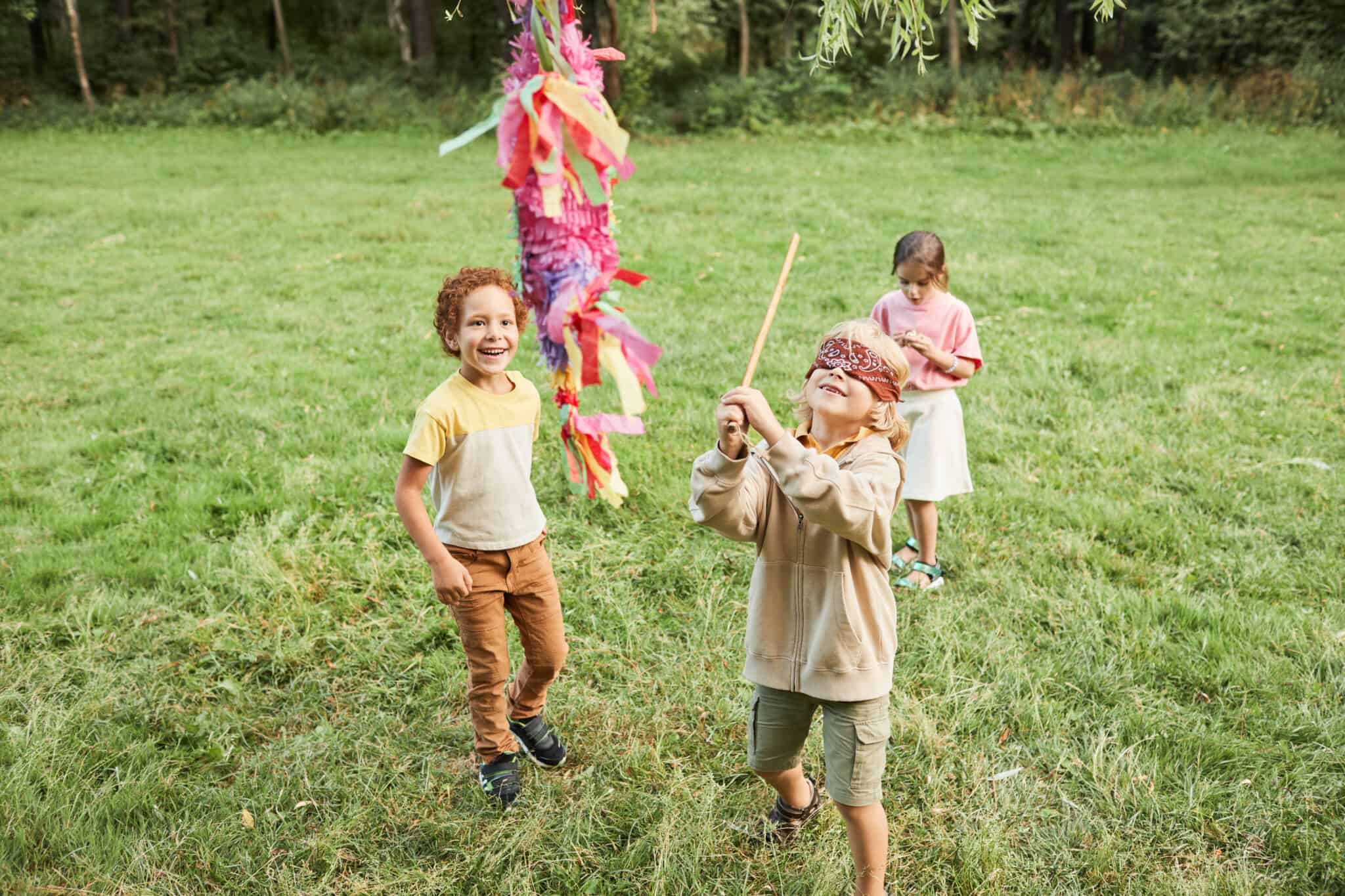 três crianças, dois meninos e uma menina, brincam num campo de relva, com uma pinhata, enquanto celebram o Dia da Criança