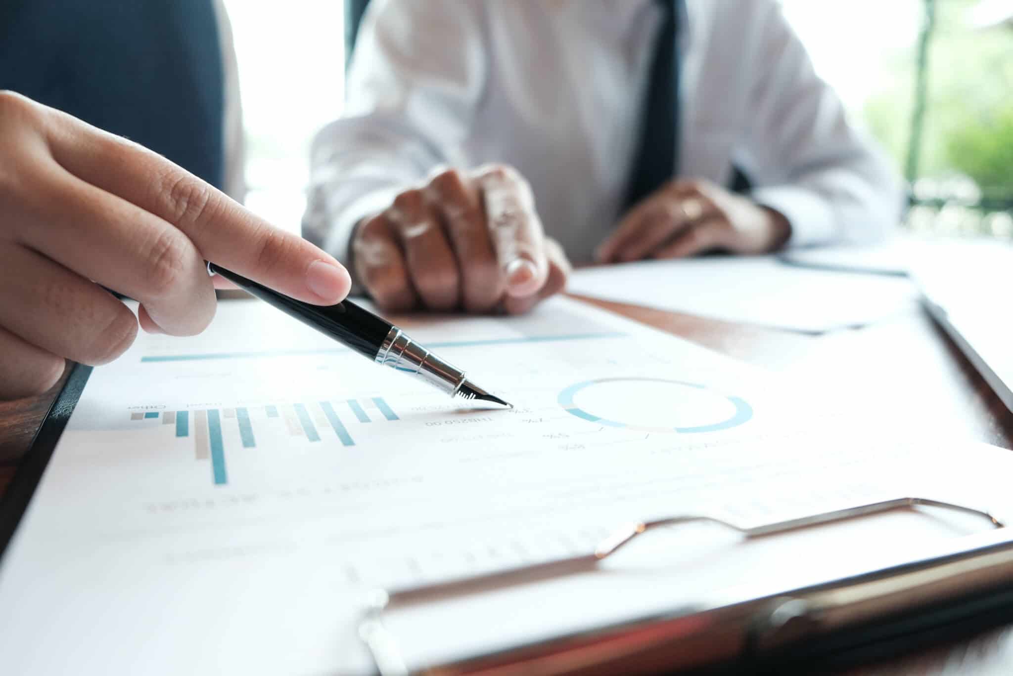 mesa cheia de documents financeiros, com gráficos, a ser revistos por dois homens, em imagem desfocada, que analisam os 10 maiores bancos do mundo