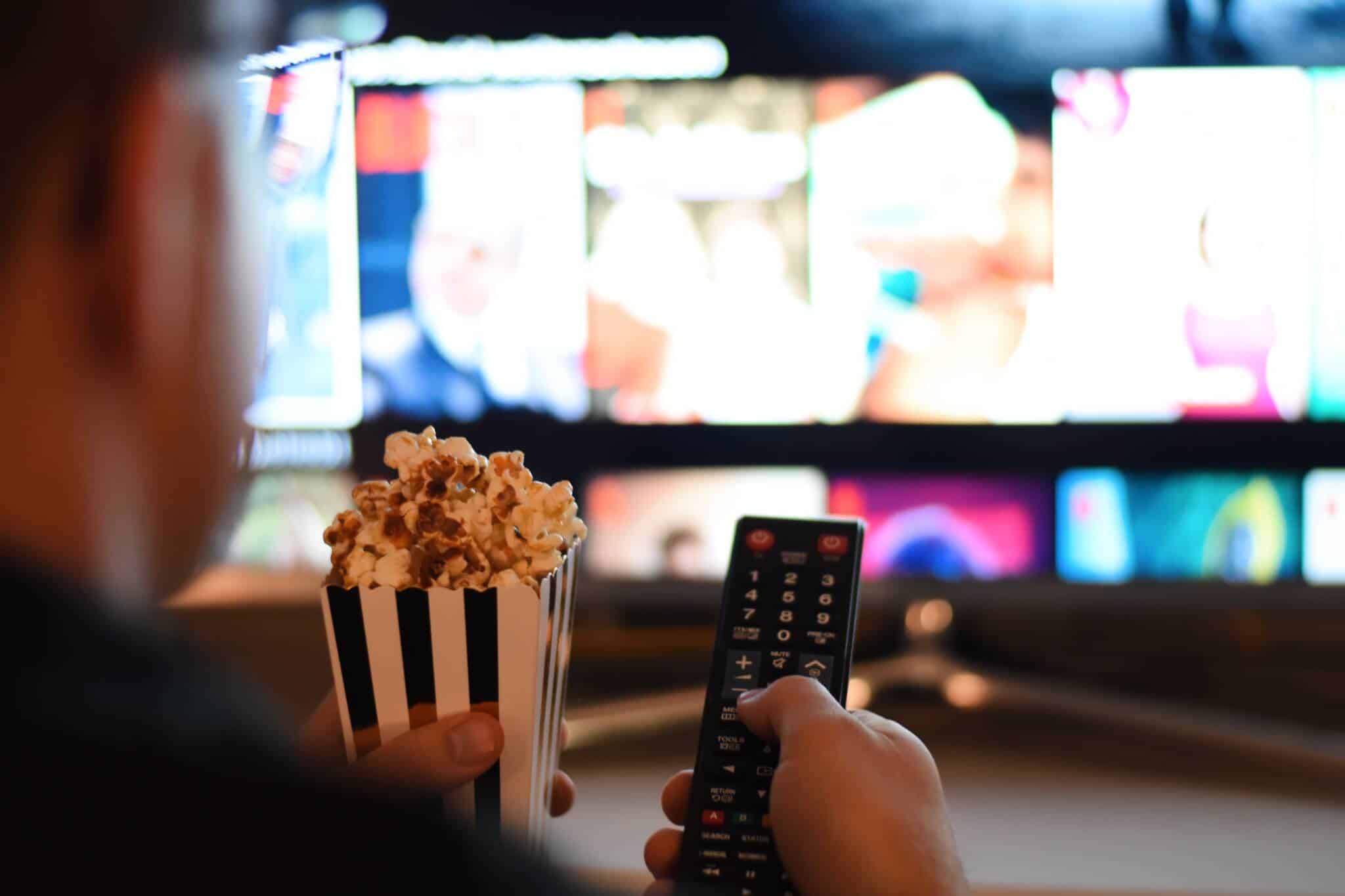 frente a uma televisão, com ecran desfocado, a mão de um jovem rapaz segura comando e pipocas, pronto para ver filmes que ajudam na educação financeira
