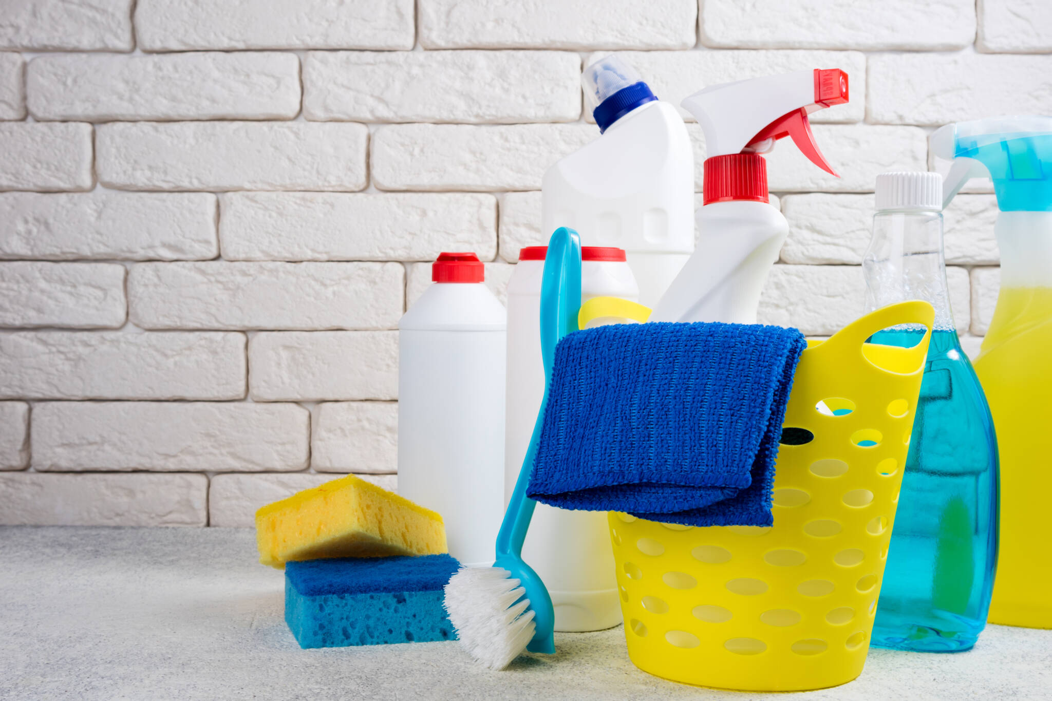 conjunto de diferentes detergentes e utensílios de limpeza em cima de bancada branca de cozinha, com fundo de azulejos brancos, remtendo para trabalho doméstico