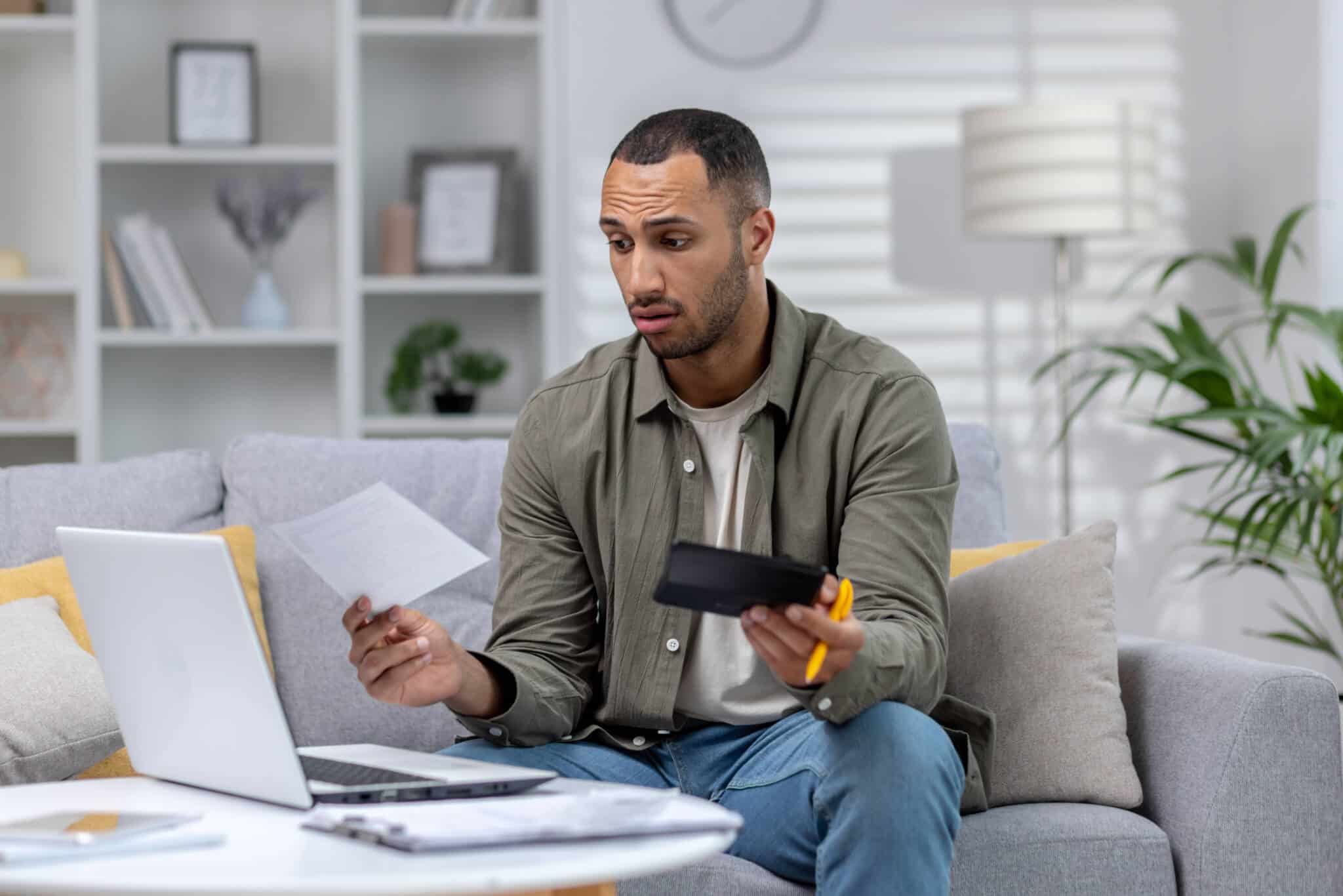 jovem homem a trabalhar na sua sala, ao computador, segura calculadora e documentos enquanto tenta perceber a gestão de responsabilidades do trabalho extra ou part-time