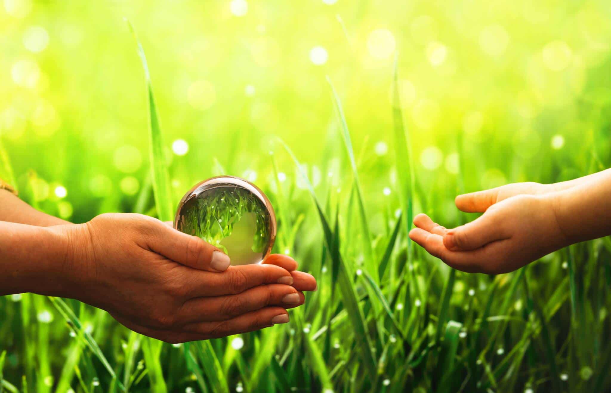 em fundo de relva verde duas maos de pequenas crianças trocam um globo de vidro transparente que ilustra o planeta, num cenário que remete para os impostos e taxas ambientais que pagamos