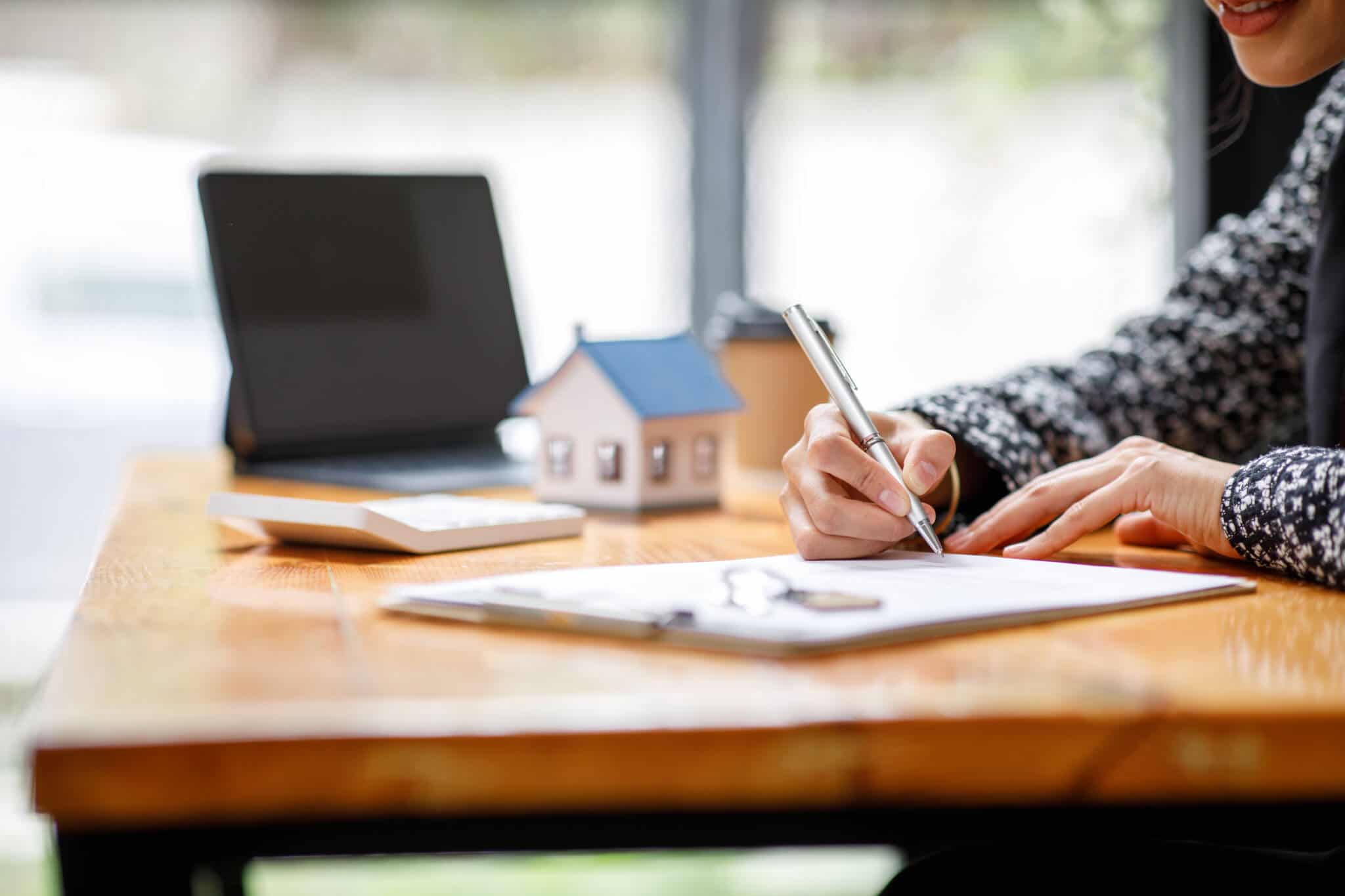 pessoa a fazer contas num papel com um portátil em cima da mesa e uma casa a ilustrar a Isenção de mais-valias para maiores de 65 anos