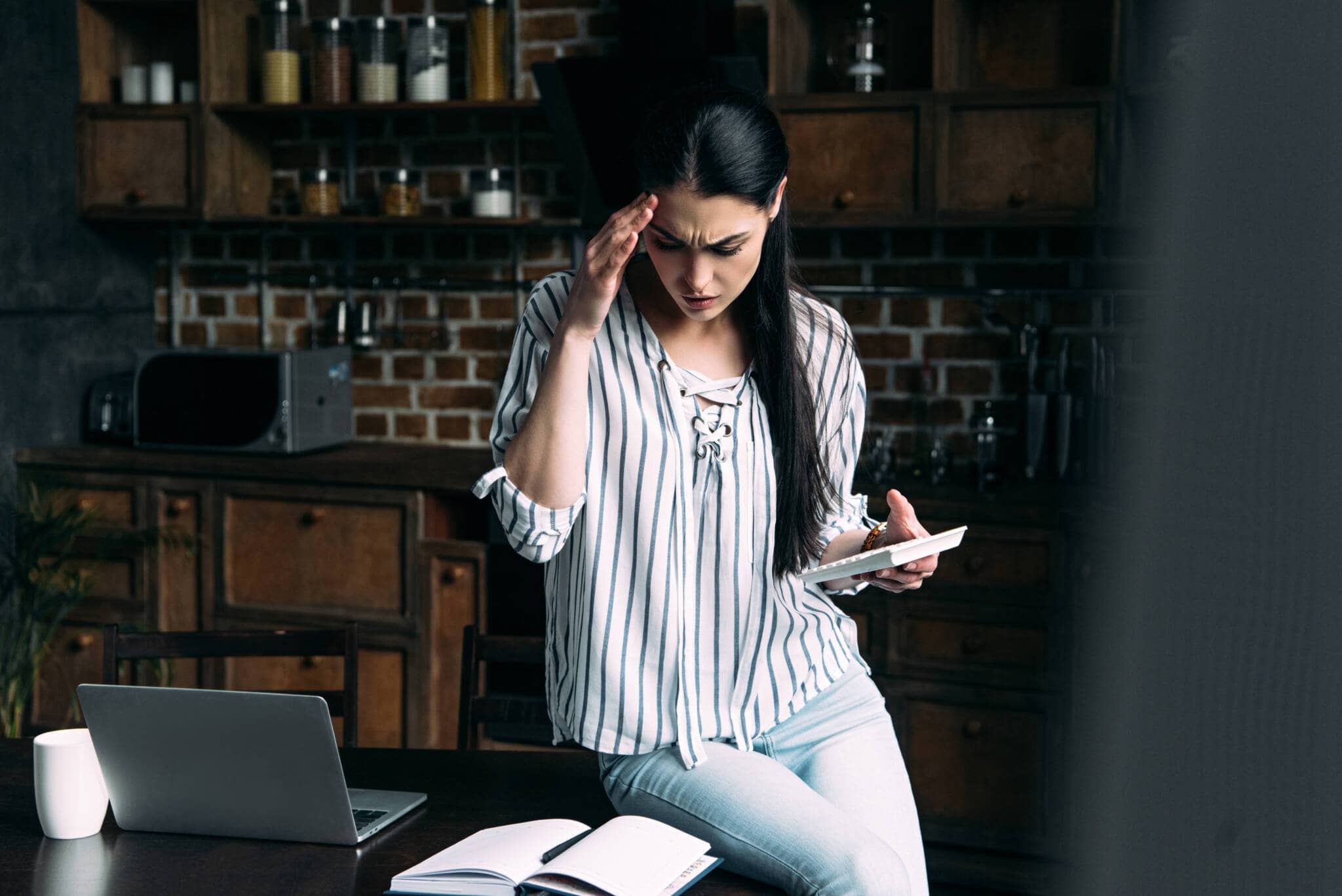 jovem mulher com um ar preocupdo, com a mão na cabeça, está em pé junto a uma mesa com cadernos e documentos e segura uma calculadora para tentar perceber o cálculo da retenção na fonte e as diferenças com o imposto