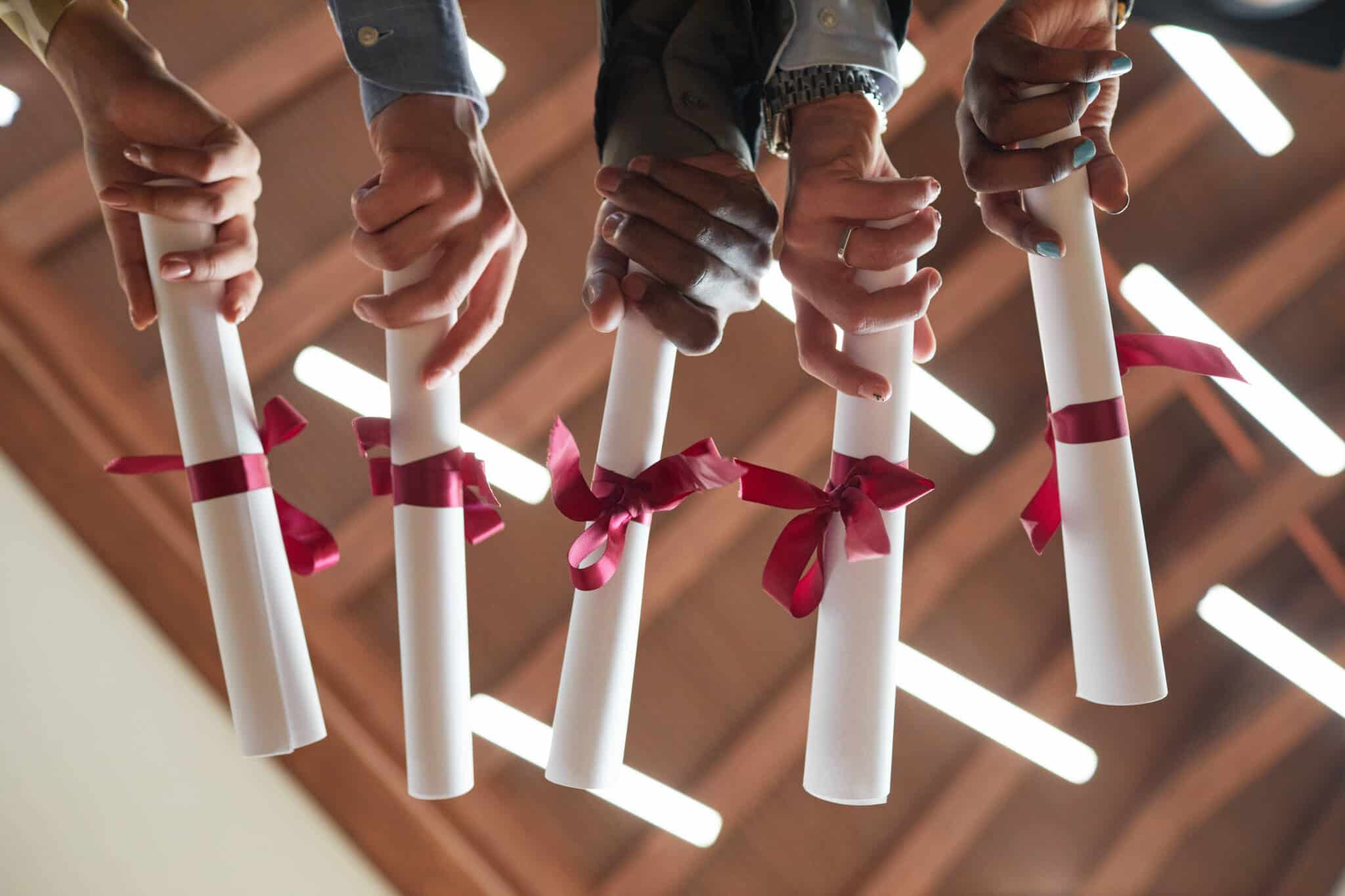 mãos de jovens, de diferentes etnias, seguram o canudo que simboliza o fim dos estudos, sendo certificado de habilitações e um diploma