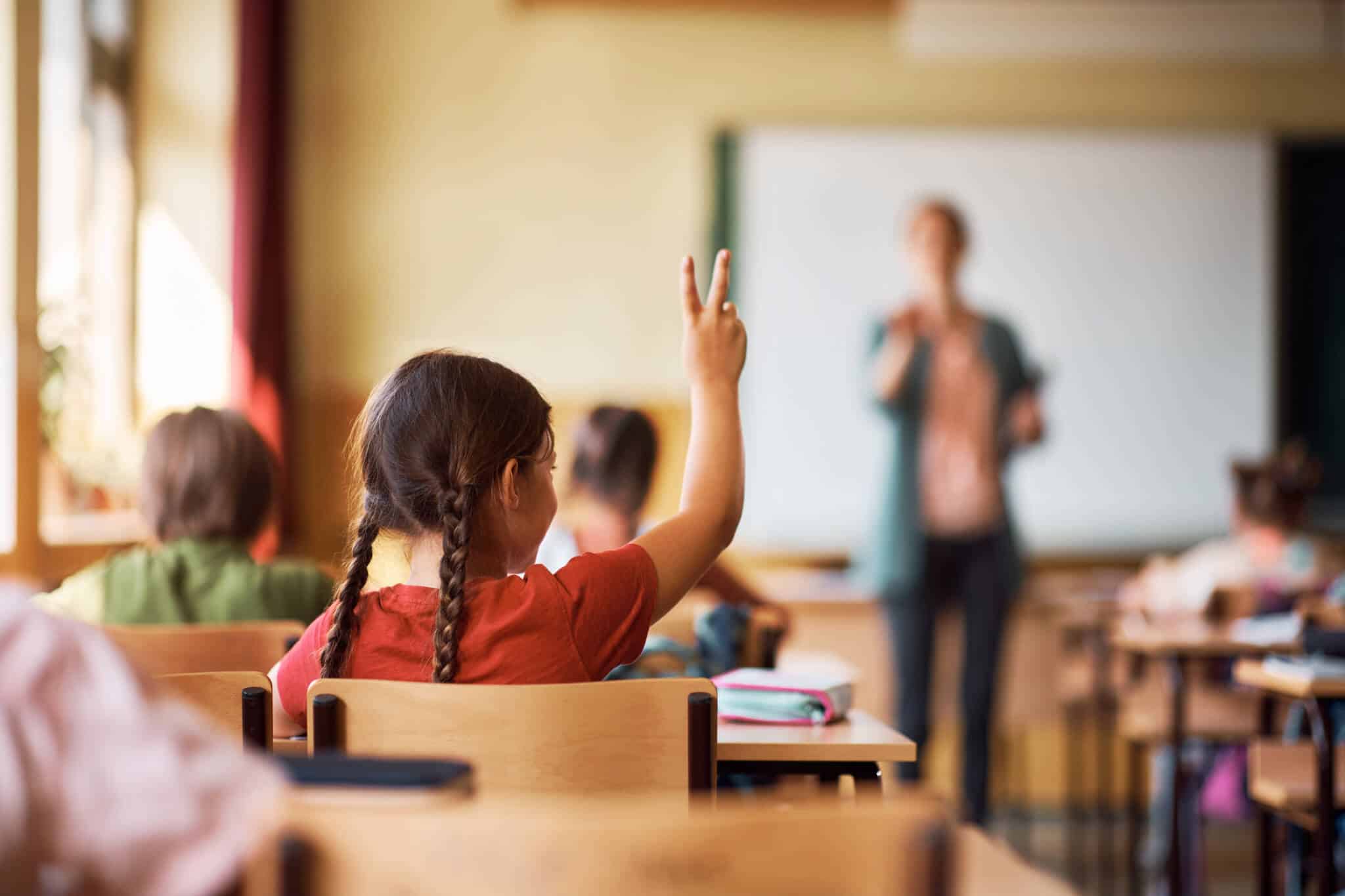 Criança com cerca de 7 anos sentada numa sala de aula, com o dedo no ar para responder à professora que se encontra ao fundo da sala.
