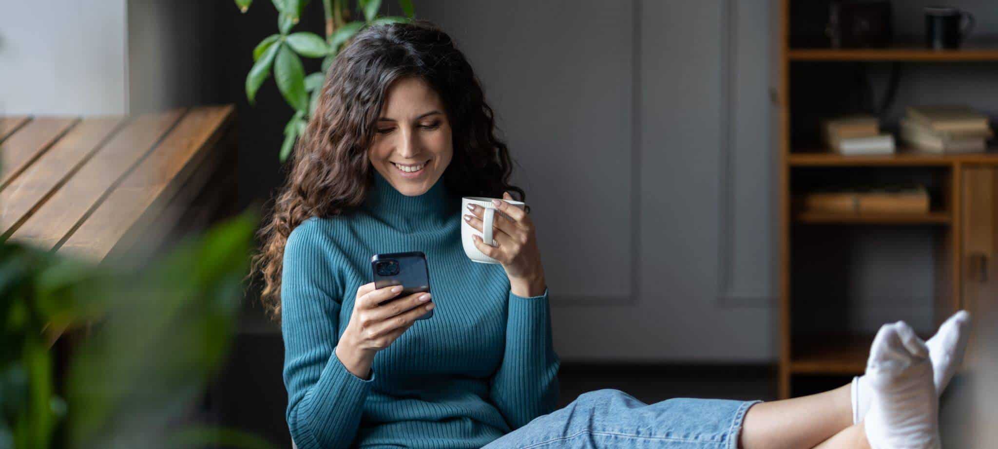 mulher sentada em frente da secretária com o telemóvel numa mão e uma caneca na outra