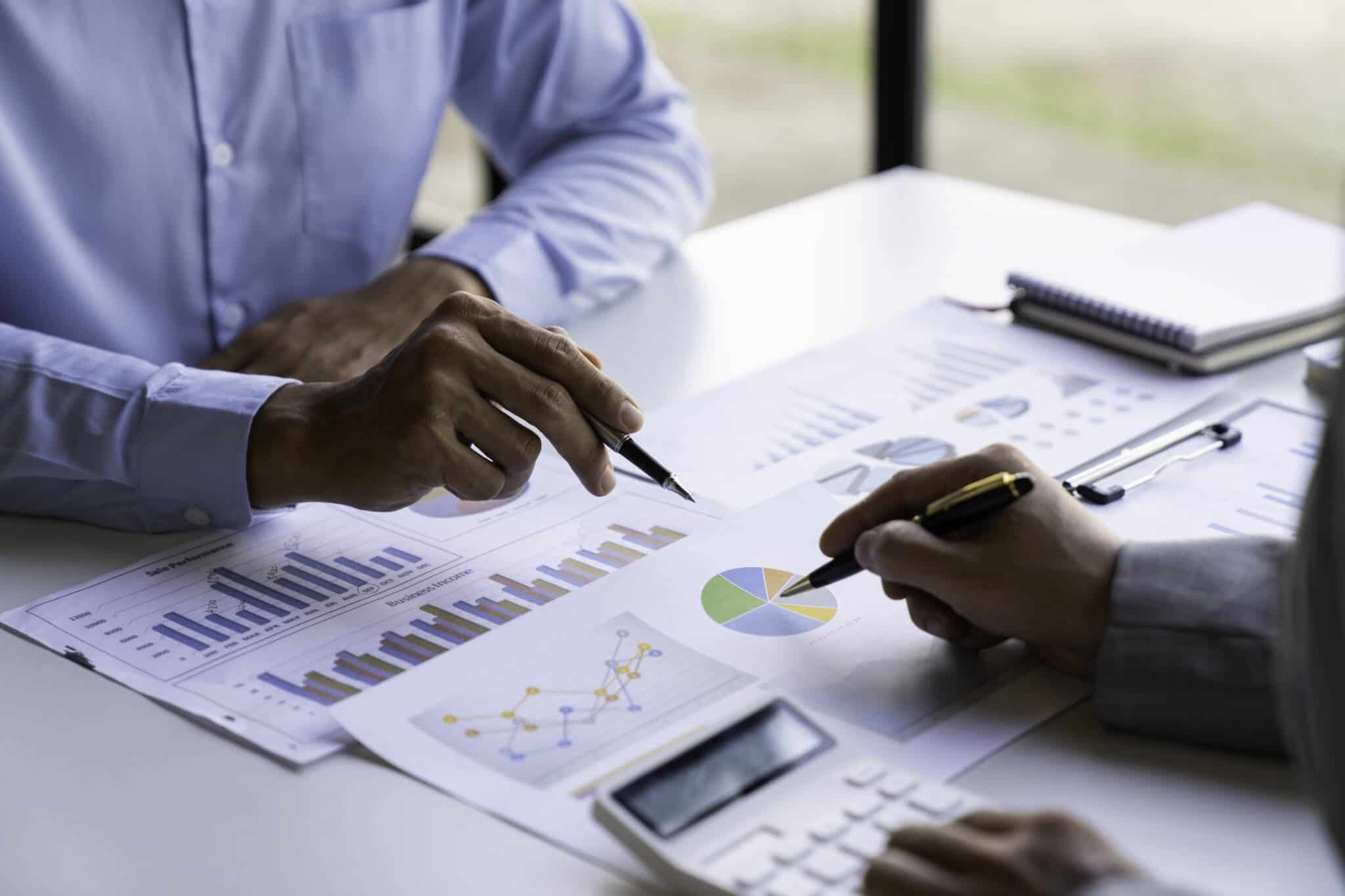 mesa coberta de documentos com gráficos e números que estão a ser calculados por dois homens com utilização da calculadora numa avaliação de empresa