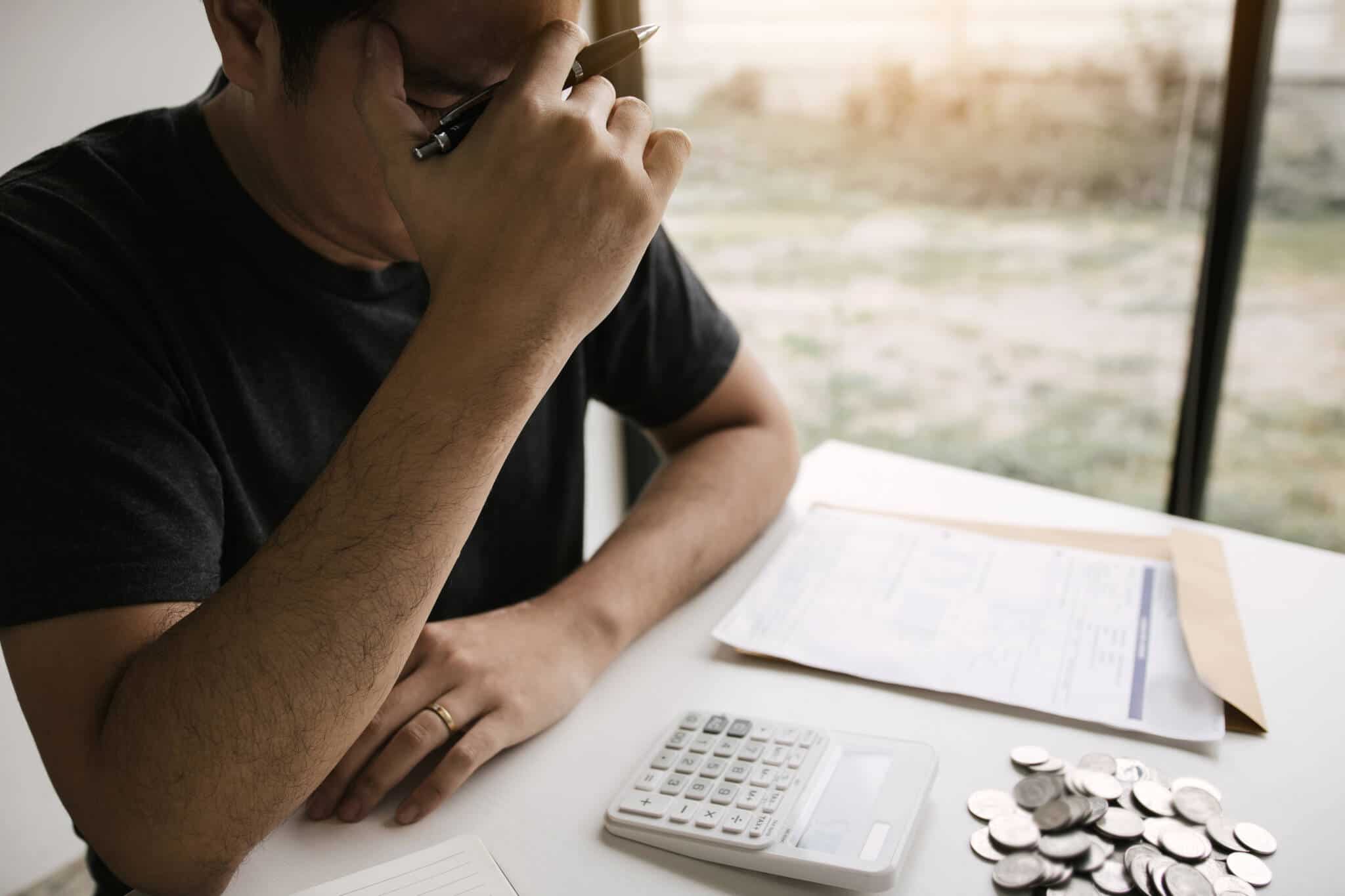 jovem rapaz a fazer contas com a calculadora, caneta e muitos papeis espalhados numa mesa, junto a uma janela, está com a mão na cabeça mostrando stress financeiro, o qual pode ser medido num simulador