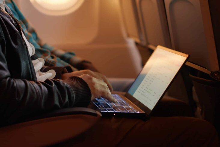 jovem homem sentado junto à hanela do avião está com o computador portátil no colo, porque vai viajar em trabalho