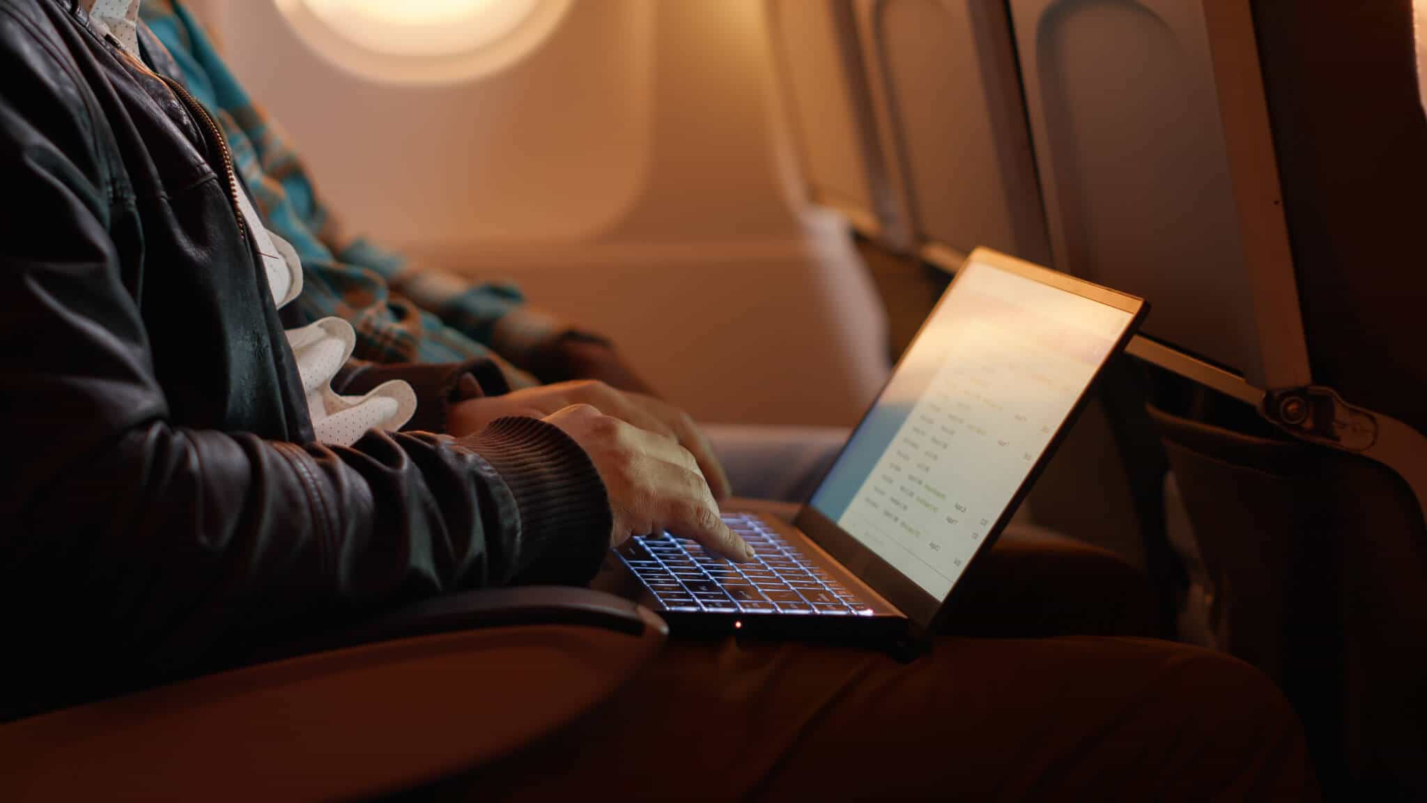 jovem homem sentado junto à hanela do avião está com o computador portátil no colo, porque vai viajar em trabalho
