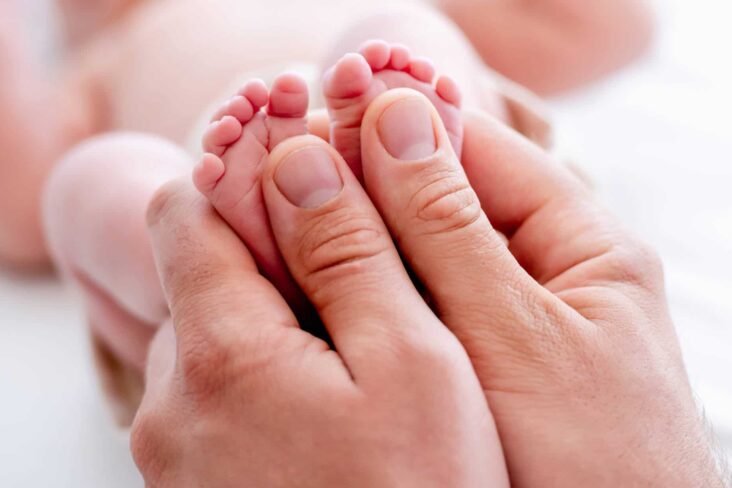 mãos agarram pés de recém nascido deitado de barriga para cima, em fundo desfocado