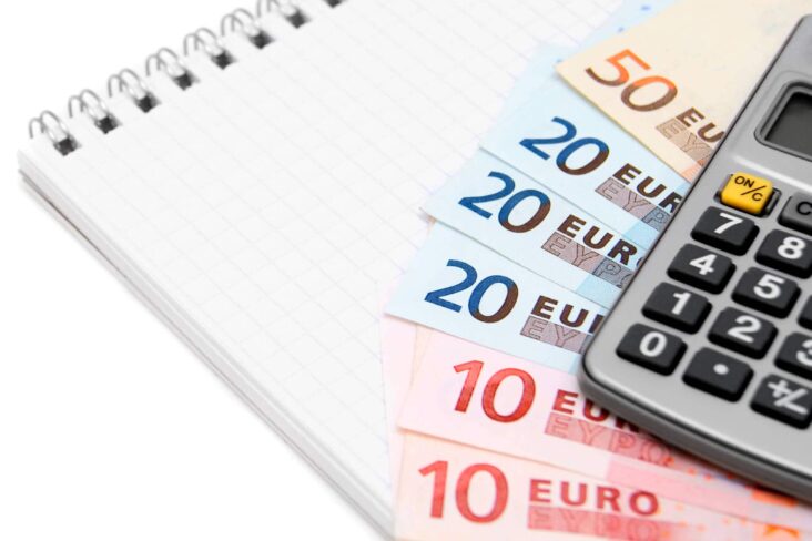 Uma série de notas de euro (50, 20 e 10 euros) colocadas em cima de um bloco de notas em formato leque. Por cima está uma calculadora, numa alusão à contabilidade