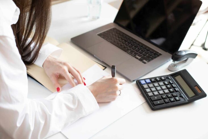 mulher a anotar os dados de uma simulação de crédito para comparar propostas, enquanto tem o portátil e a calculadora em cima da mesa