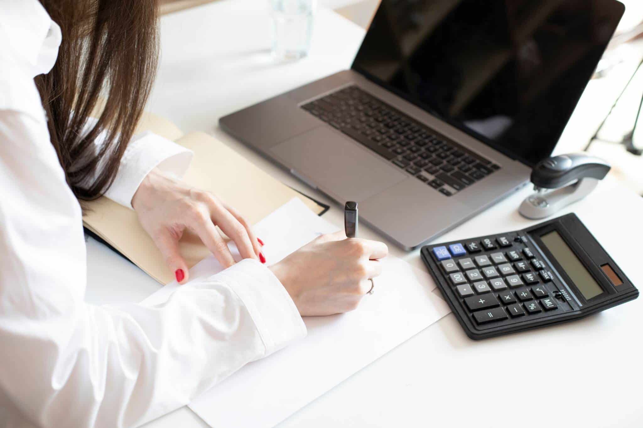 mulher a anotar os dados de uma simulação de crédito para comparar propostas, enquanto tem o portátil e a calculadora em cima da mesa