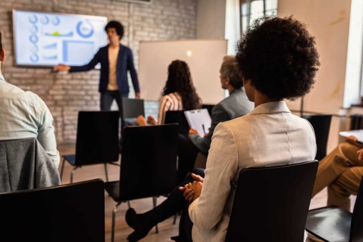 jovem rapaz em fundo desfocado, dá formação a um conjunto de jovens sentados em cadeiras e mesas individuais, e para tal obteve certificação de formadores