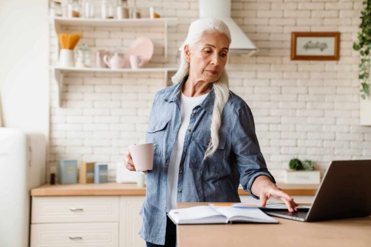 mulher sénior, de longo cabelo branco, a trabalhar em casa no seu portátil e em ambiente de cozinha, o que lhe é permitido trabalhar na reforma