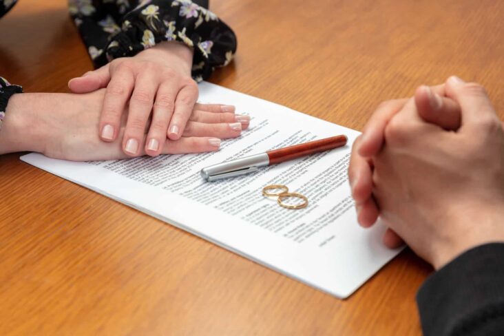 mãos de um casal frente a frente, sob um documento , junto a umas alianças, e com uma caneta para assinar após a decisão de escolher entre a união de facto e casamento