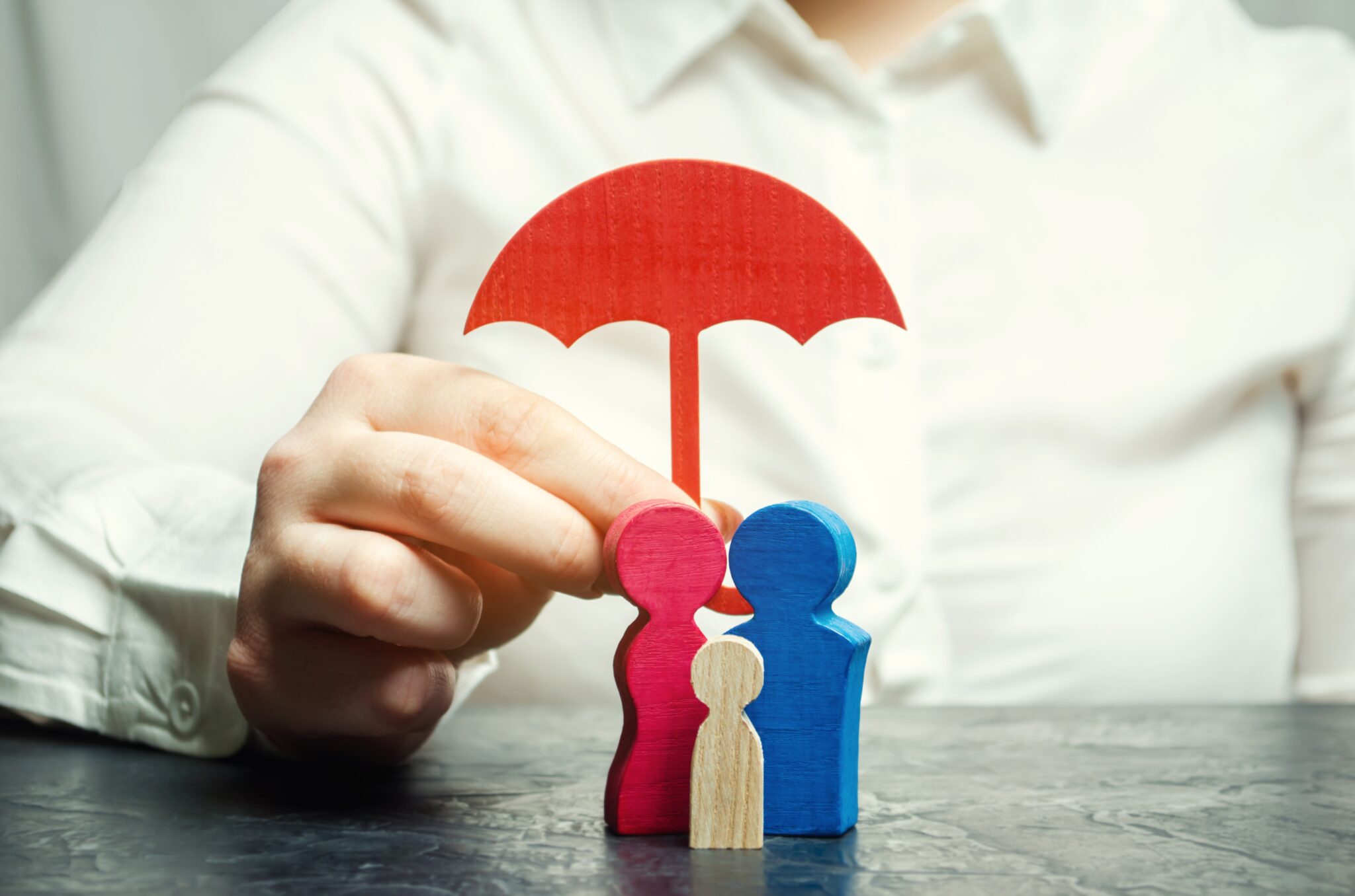 mão de homem, desfocado no fundo da imagem, segura um chapéu de chuva vermelho que está a proteger 3 pões de madeira e diferentes cores que simbolizam mãe, pai e filho, abrangidos por um seguro com período de carência