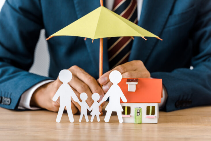 seguro multirriscos ilustrado por produção fotográfica com figuras de papel que são uma família, bem como a fachada de uma casa sob um chapéu de chuva que protege segurado por um executivo