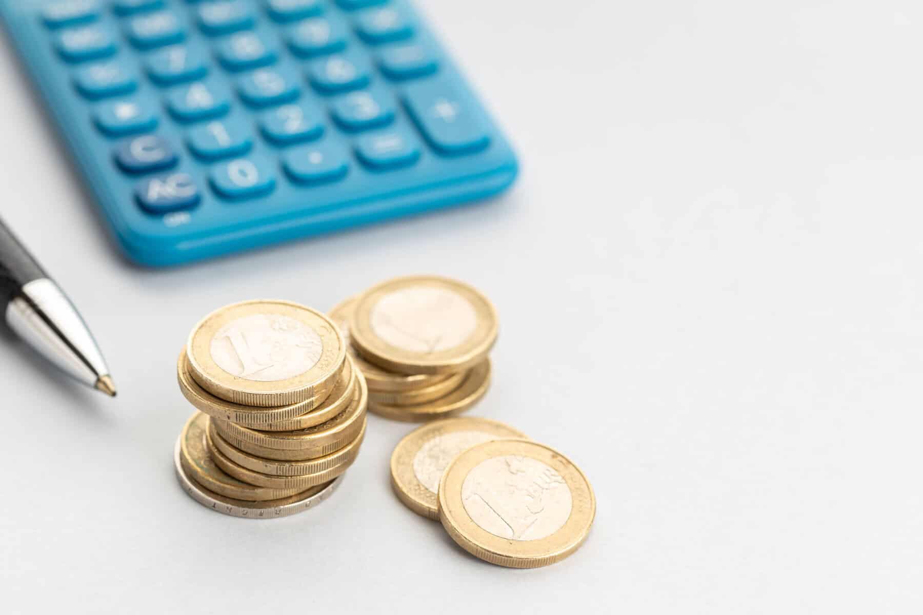 Três molhos de moedas de euro sobre uma mesa, ao lado de uma calculadora azul e de uma caneta, numa alusão a investimentos com pouco dinheiro