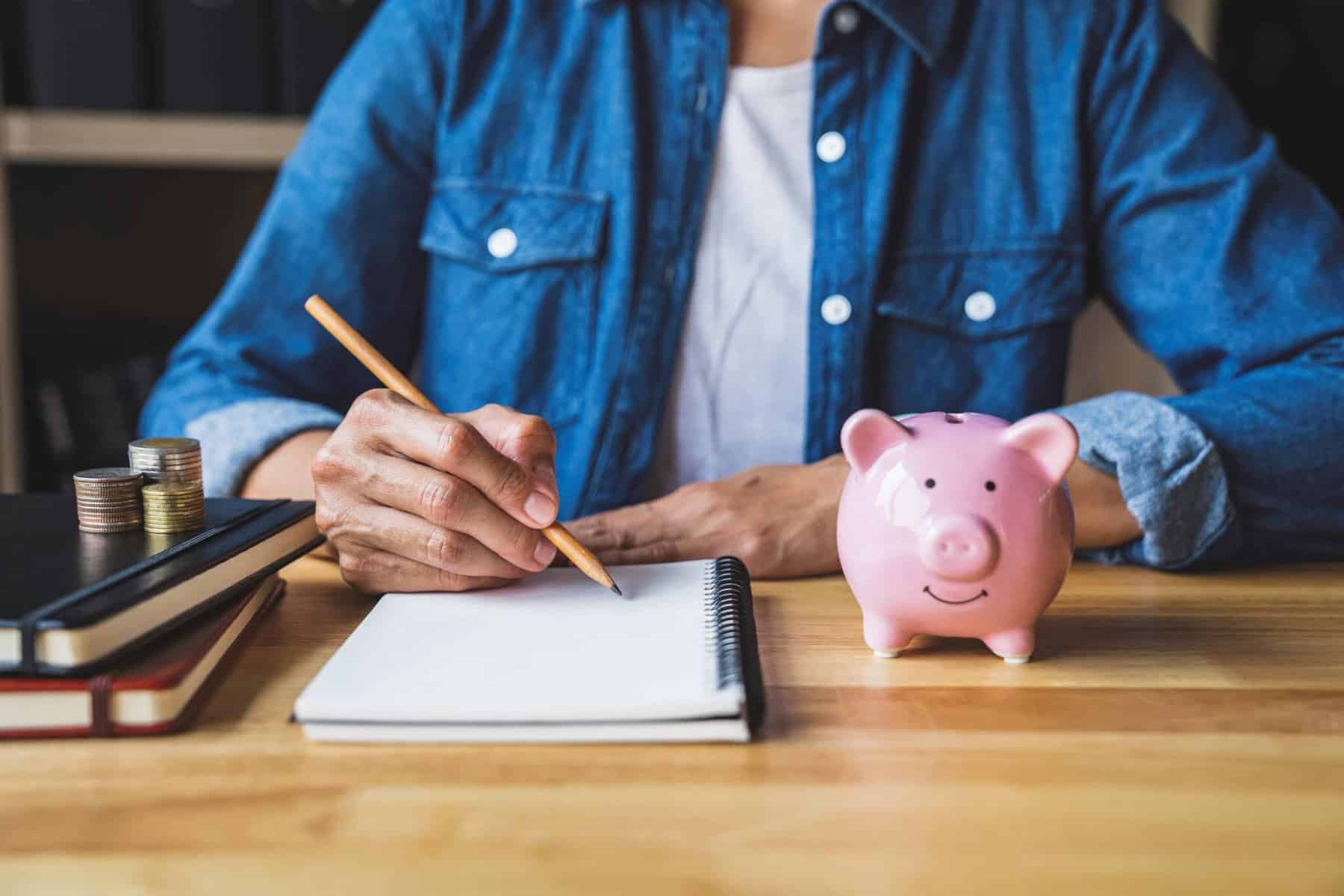 pessoa com um caderno e um lápis a escolher ppr. Ao lado, um porco mealheiro
