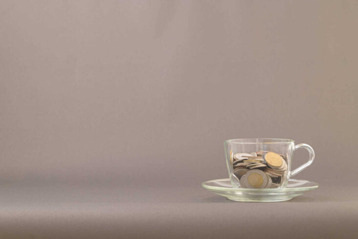 chavena de café de vidro transparente pousada em cima de um pires sobre uma mesa cinzenta. Dentro da chávena estão moedas de euro, numa alusão aos primeiros passos para começar a investir