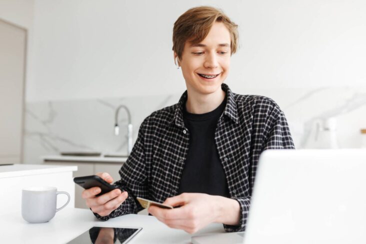 jovem sorridente com um cartão bancário na mão a olhar para o computador a ilustrar como abrir uma conta para um filho