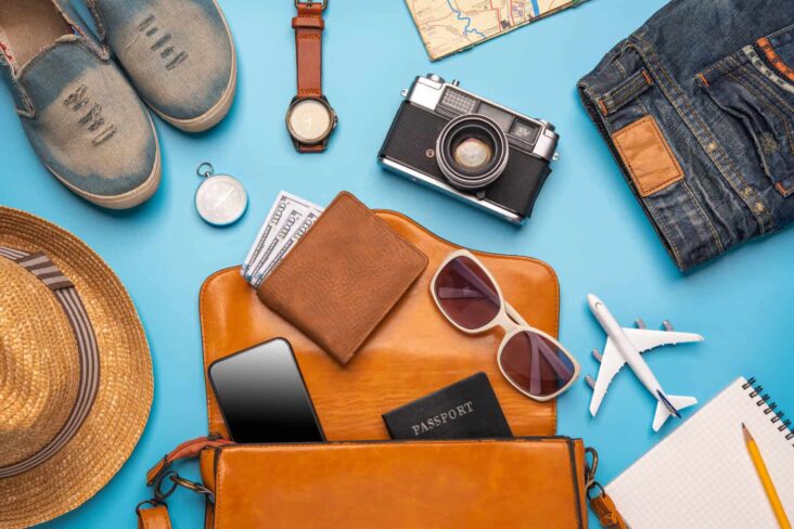 carteira, óculos de sol, máquina fotográfica, roupa e sapatos, relógio e o passaporte espalhados em cima de uma mesa azul, numa alusão a uma viagem internacional