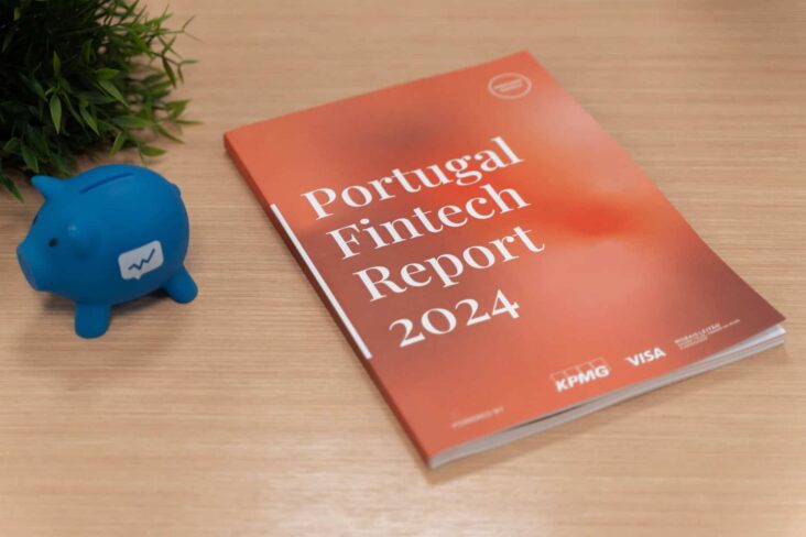 Relatório do Portugal Fintech de 2024, onde o Doutor Finanças surge pelo sétimo ano. O relatório, que está em formato de livro, está pousado em cima de uma mesa de madeira. Ao seu lado está pousado um mealheiro azul com o logo do Doutor Finanças.
