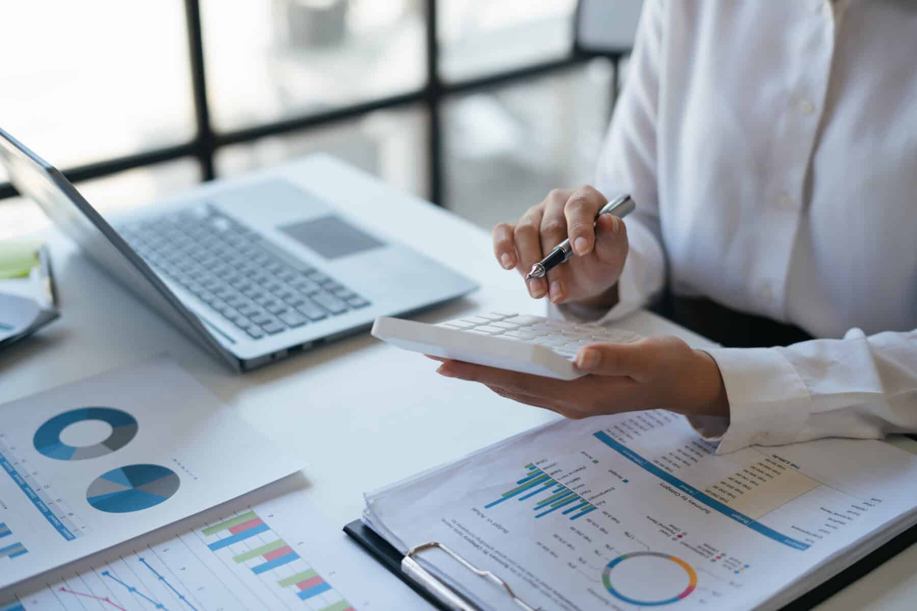Mãos de mulher seguram uma máquina de calcular. Em frente tem o computador e várias folhas com gráficos e informação sobre o inventário para enviar para a Autoridade Tributária