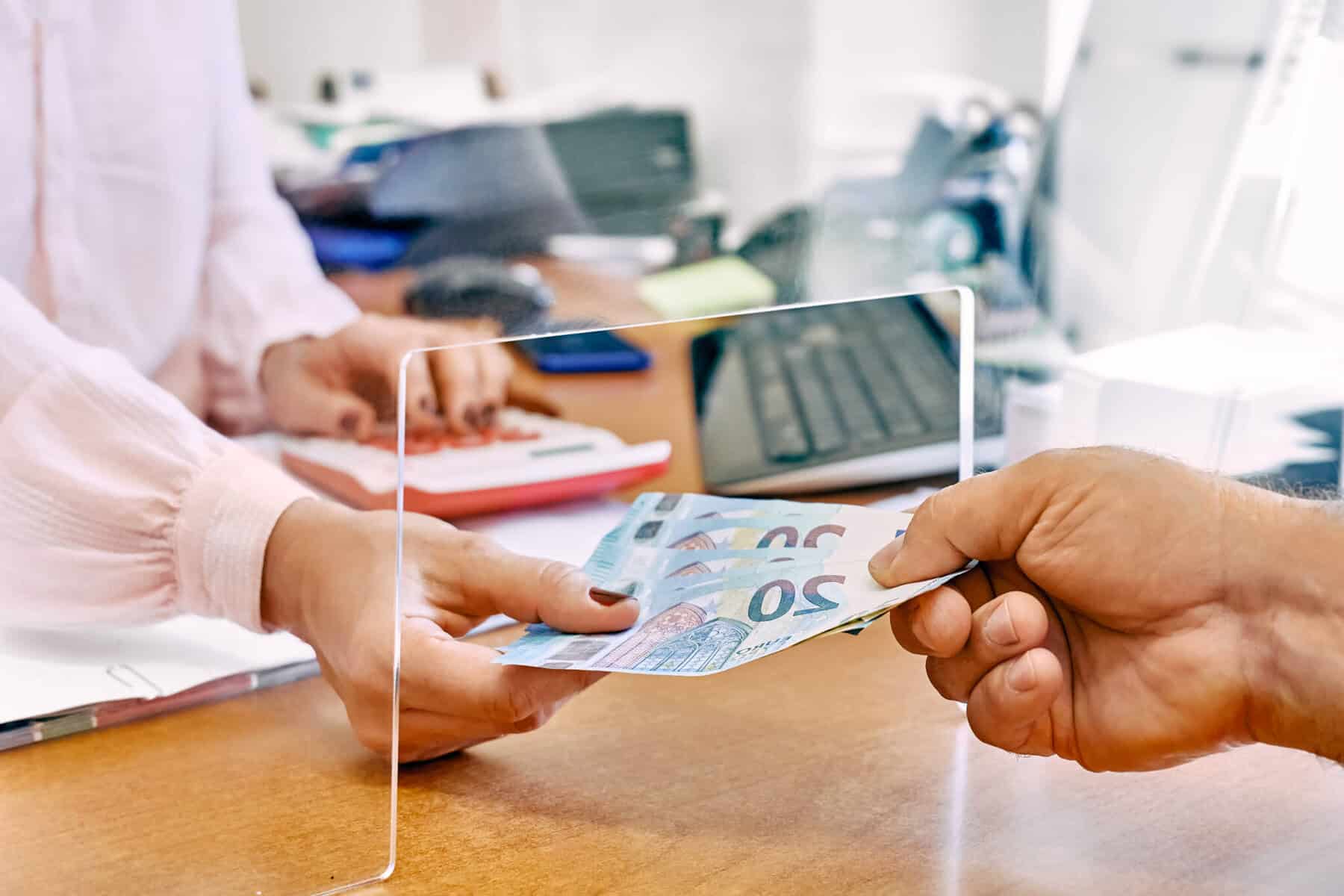 Mão de homem entrega notas de 20 euros a uma mão de mulher, que está dentro de um guichê de atendimento público