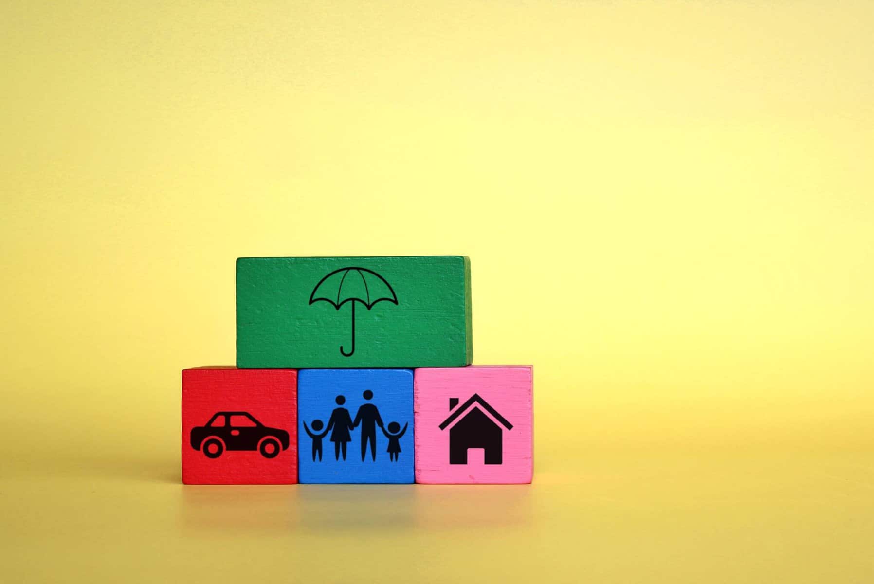 Três cubos coloridos com desenhos de um carro, uma família e uma casa. Por cima, está uma quarto cubo com um chapéu de chuva desenhado, numa alusão aos seguros de vida