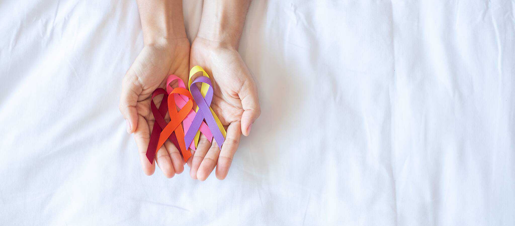 Duas mãos de mulher juntas, em formato de concha, seguram laços de várias cores, numa alusão ao cancro