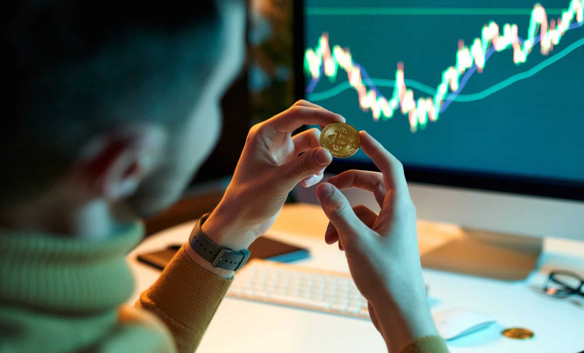 Investidor jovem olha para símbolo físico da bitcoin em frente a um computador com um gráfico de mercados financeiros.