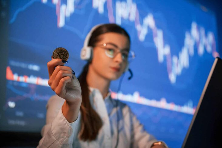 Uma tarder segura um símbolo físico da bitcoin na mão, com um ecrã gigante atrás com um gráfico, de auriculares nos ouvidos e um computador portátil à sua frente.