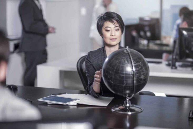 Mulher asiática olha para um globo à secretária, com folhas com contas na mesa.