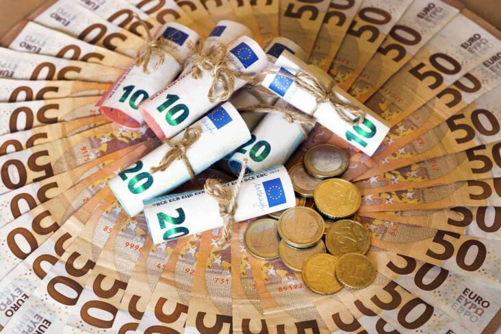 Várias notas de dólar dispostas sobre uma mesa. Ao centro rolos de notas e moedas de euro