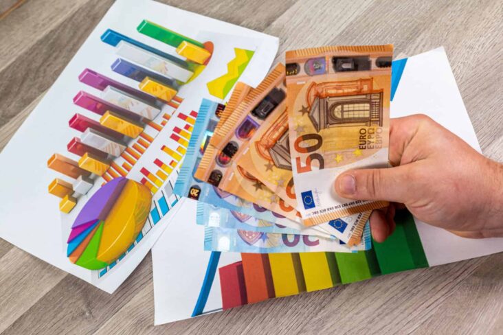 Homem segura várias notas de 50 euros. Pousadas na mesa estão algumas folhas com gráficos de análise financeira