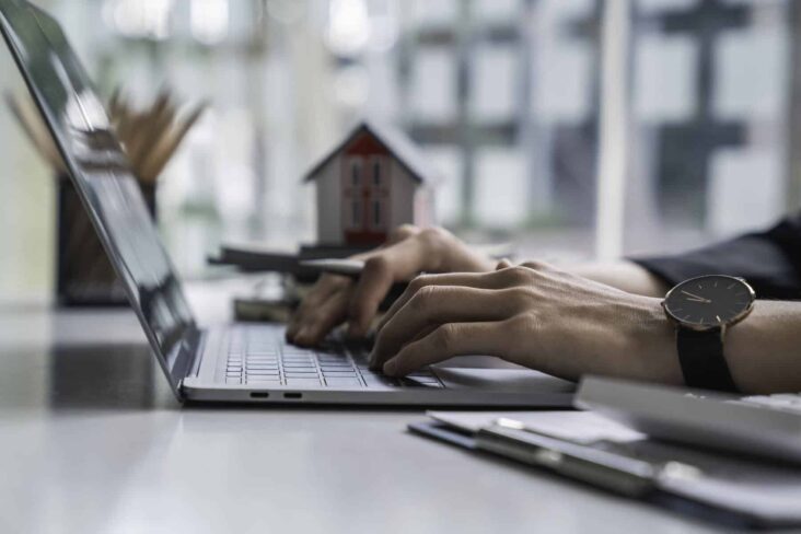 mãos a teclar num computador com uma casa ao fundo da imagem a ilustrar o tema Sites e apps para procurar casa