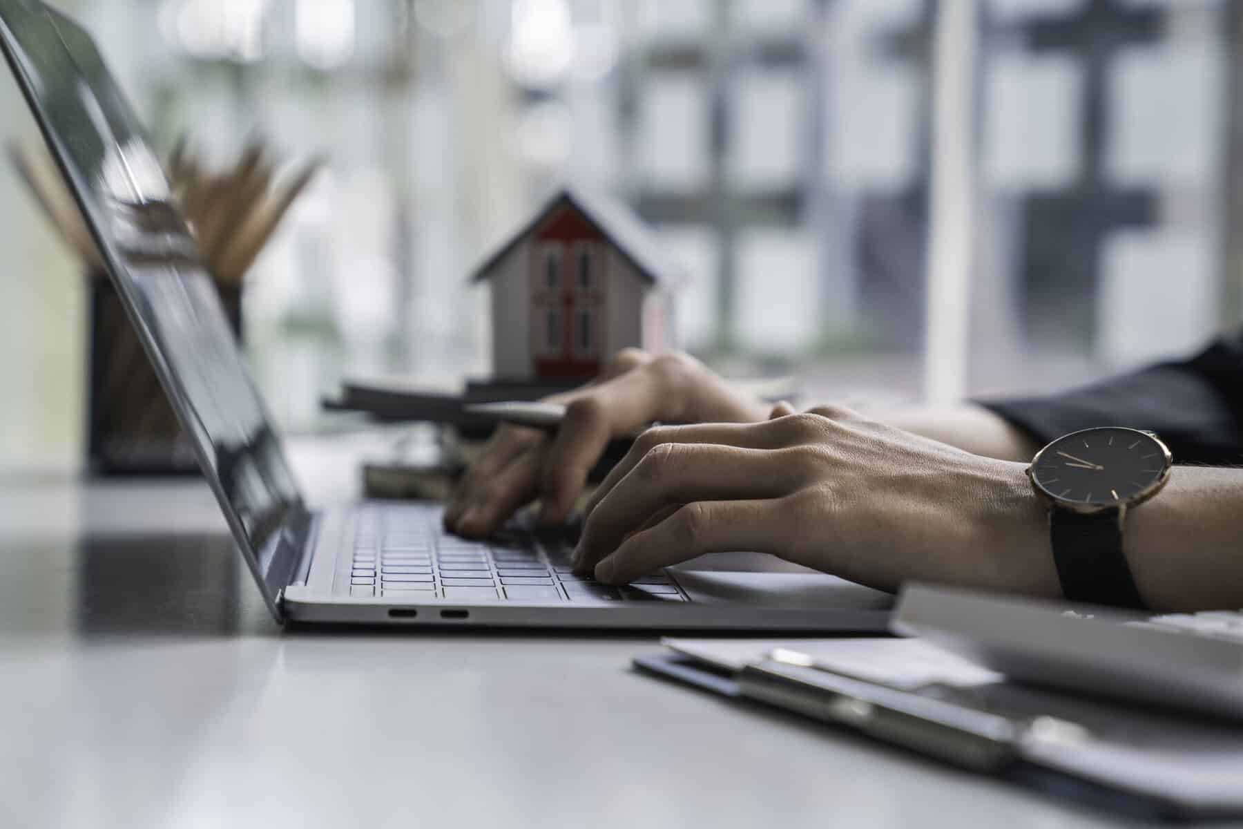 mãos a teclar num computador com uma casa ao fundo da imagem a ilustrar o tema Sites e apps para procurar casa