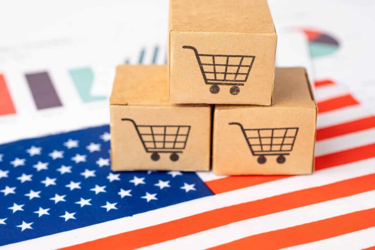 Bandeira dos EUA exposta em cima de uma mesa, Por cima estão três cubos com a imagem de carrinhos de compras, numa alusão às tarifas comerciais anunciadas por Trump